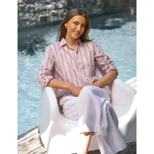 Woman sitting by a pool wearing a pink striped shirt and white pants.