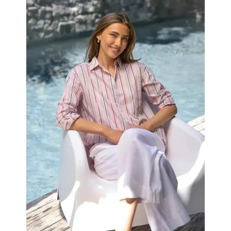 Woman sitting by a pool wearing a pink striped shirt and white pants.